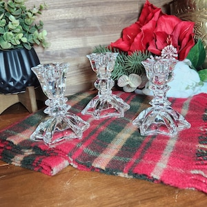 Puede incluir: Tres candelabros de cristal transparente con base de estrella están dispuestos sobre una tela a cuadros roja y verde. Los candelabros tienen un diseño facetado y se exhiben sobre una superficie de madera. Hay objetos decorativos en el fondo.