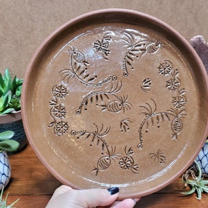 May include: A brown ceramic plate with a raised rim. The plate features an etched design of stylized fish, floral, and geometric shapes. The plate is round and appears to be handmade.