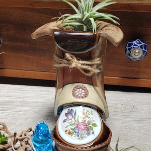 May include: A unique, handcrafted ceramic air plant holder in shades of brown, tan, and cream, adorned with a floral design. The holder is tied with twine and features a decorative floral plate at the base. A small air plant sits inside.