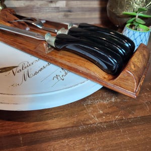 May include: A wooden serving tray holding a set of black-handled knives and a carving fork. The tray rests on a white plate with script writing. The knife handles are curved and glossy.