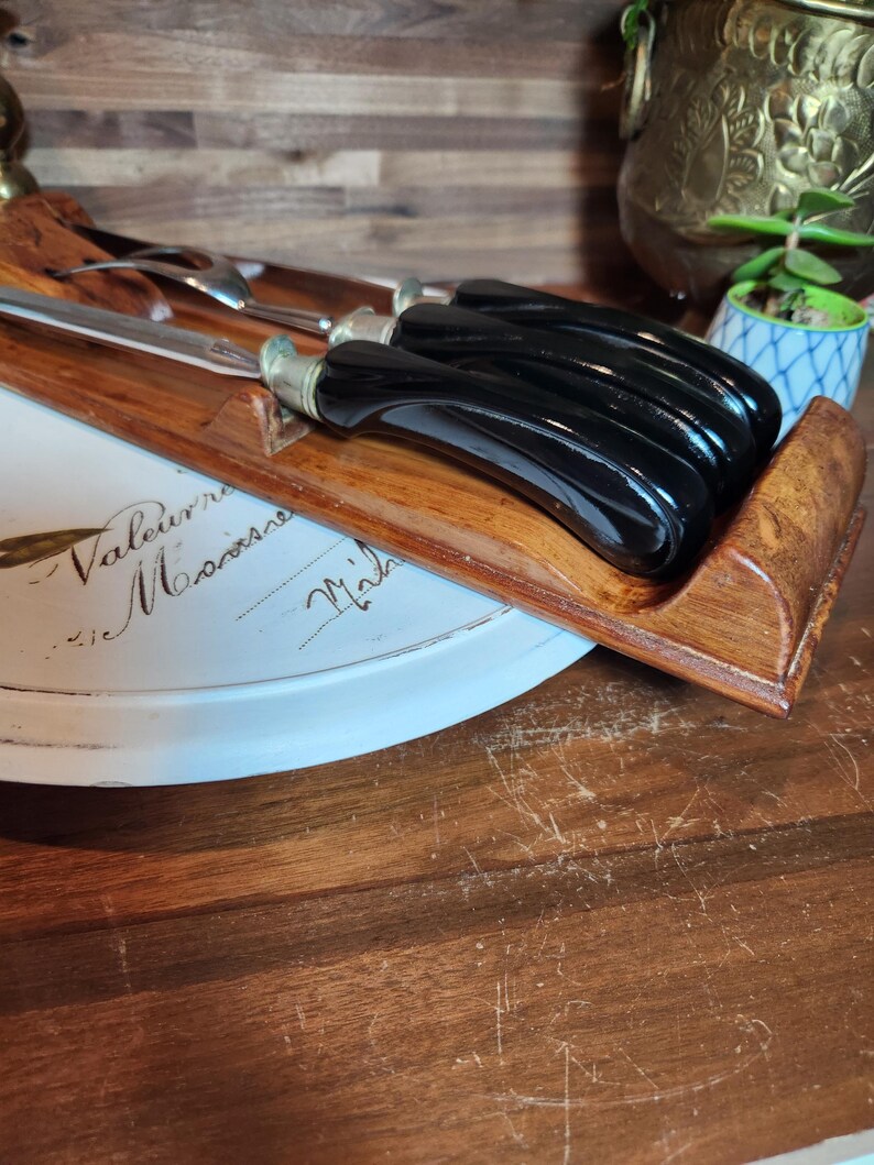 May include: A wooden serving tray holding a set of black-handled knives and a carving fork. The tray rests on a white plate with script writing. The knife handles are curved and glossy.