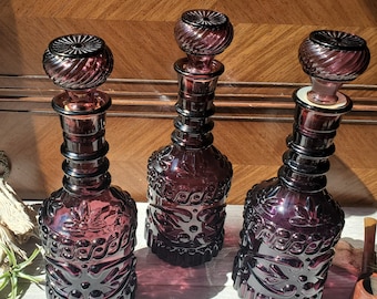 May include: Three antique, dark purple glass decanters with ornate floral and swirl designs. Each decanter has a matching stopper. The bottles are displayed on a light-colored surface with a wooden background.