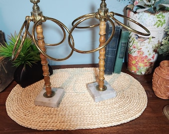Può includere: Due portasciugamani vintage. Ogni anello presenta una finitura in ottone, un palo in legno chiaro e una base quadrata in marmo. Gli anelli sono disposti su un tappetino ovale intrecciato, con oggetti decorativi sullo sfondo.