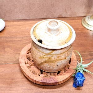 May include: A tan ceramic sugar bowl with a lid and a small spoon, resting on a carved wooden coaster. The bowl has brown streaks and a small handle on the lid. A small blue elephant figurine is next to the coaster.