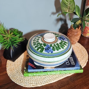 May include: A ceramic lidded bowl with a floral design in blue and green. The bowl sits atop two cookbooks on a woven placemat. The lid has a white knob and is decorated with blue flowers and green leaves.