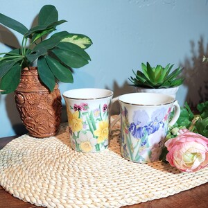 May include: Two white ceramic mugs with floral designs. One mug features yellow daffodils and pink flowers, the other has purple irises. Both have gold rims and handles. The mugs are on a woven mat.