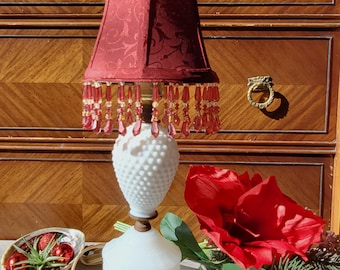 May include: A vintage table lamp with a white milk glass base and a burgundy shade. The shade is adorned with beaded fringe. A red flower and decorative tray with ornaments are also in the image. The lamp is on a wooden surface.
