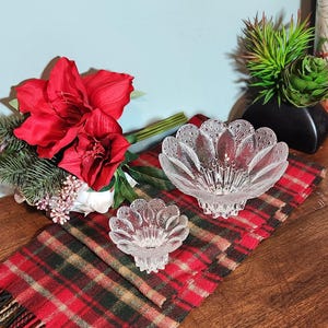 May include: Two clear, decorative glass bowls with a floral design sit on a red and green plaid scarf. A bouquet of red flowers and greenery is to the left, and a black vase with plants is in the background.