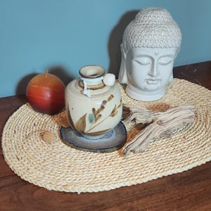 May include: A still life arrangement featuring a small, beige ceramic vase with brown and blue floral designs, a white Buddha head, a round orange candle, and a piece of driftwood. The items are arranged on a woven mat.