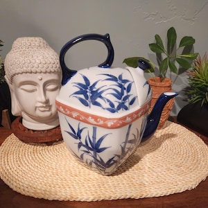 Puede incluir: Una tetera de cerámica blanca con asa y boquilla azules, decorada con hojas de bambú azules y un borde color coral. La tetera está sobre una estera tejida, junto a una estatua de cabeza de Buda blanca.