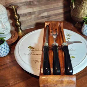 May include: A wooden cheese knife set comprising a knife, a fork, and a spreader. The black-handled utensils are displayed in a wooden holder. The set rests on a white serving tray with text.