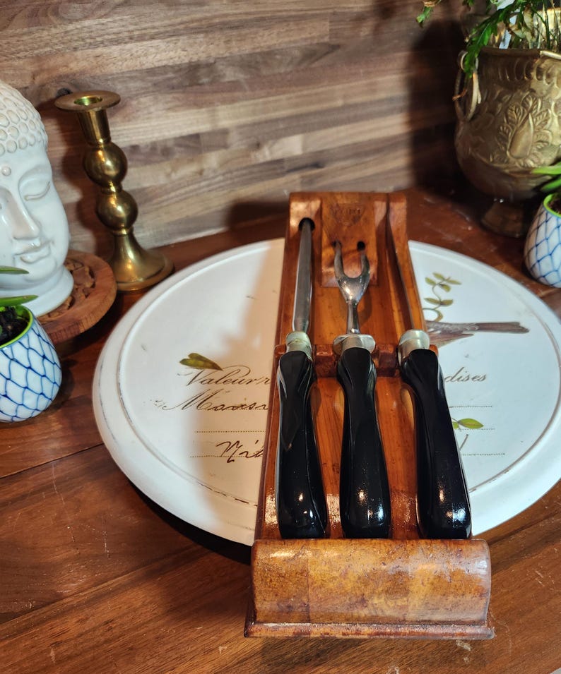 May include: A wooden cheese knife set comprising a knife, a fork, and a spreader. The black-handled utensils are displayed in a wooden holder. The set rests on a white serving tray with text.