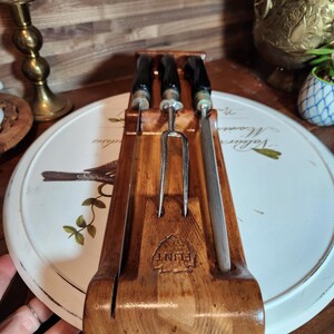 May include: A wooden carving set with a carving fork and two knives. The handles are black, and the blades are silver. The set is displayed in a wooden holder with the word "FLINT" stamped on the front.