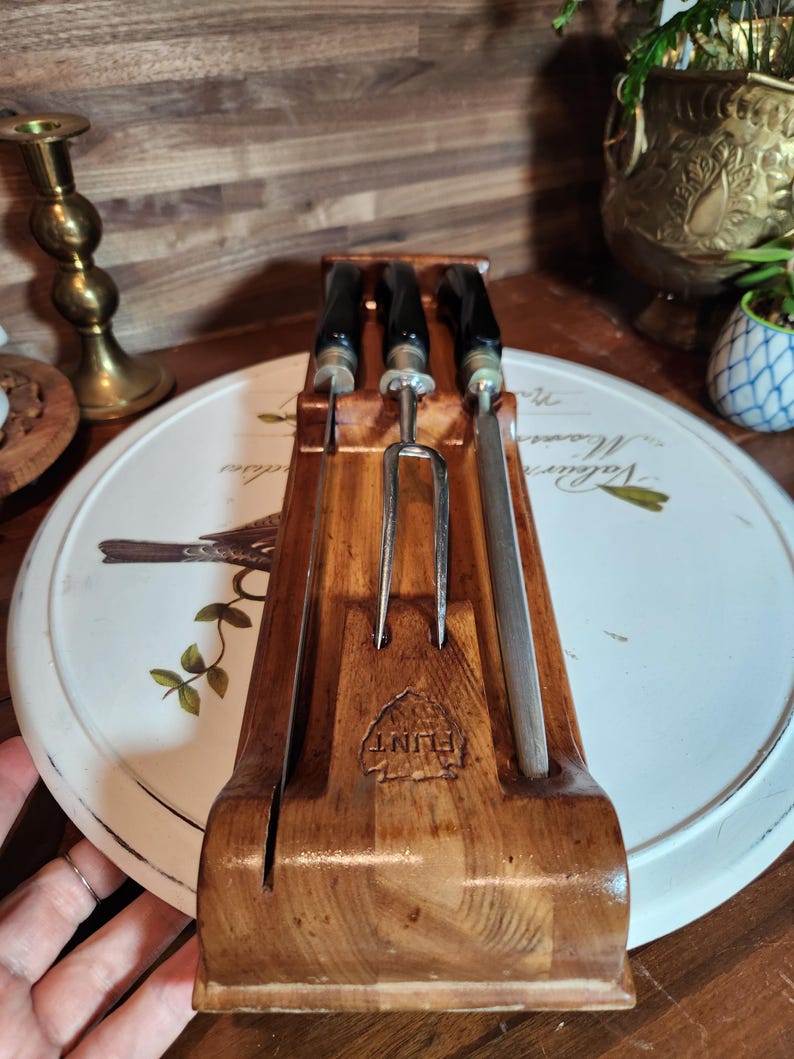 May include: A wooden carving set with a carving fork and two knives. The handles are black, and the blades are silver. The set is displayed in a wooden holder with the word "FLINT" stamped on the front.