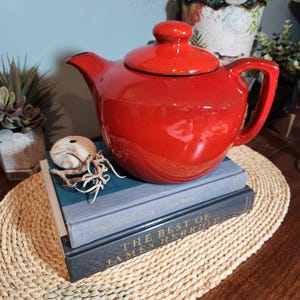 May include: A glossy red ceramic teapot with a matching lid sits atop two books. The books are stacked on a woven placemat. The teapot has a curved spout and a rounded handle. A decorative seashell is next to the teapot.