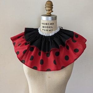 May include: A red and black ruffled collar with white lace trim and black polka dots. The collar is on a white mannequin bust.