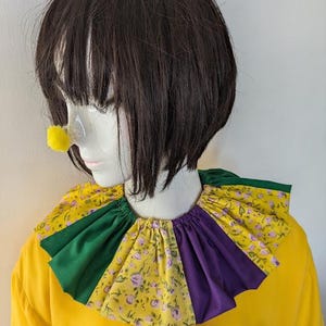 May include: A yellow clown costume with a ruffled collar featuring green, purple, and floral fabric. A yellow pom-pom nose is attached to a mannequin head with a dark brown wig. The costume is a bright, cheerful design.