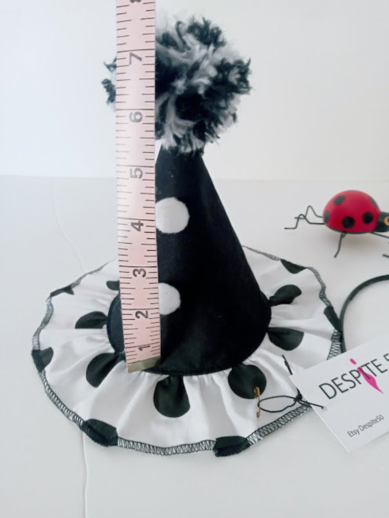 K&ouml;nnte beinhalten: Schwarz-wei&szlig;er Polka-Dot-Partyhut mit einem wei&szlig;en Bommel oben. Der Hut ist 15 cm hoch und hat einen schwarz-wei&szlig;en R&uuml;schenbesatz. Der Hut befindet sich auf einer wei&szlig;en Oberfl&auml;che mit einem Ma&szlig;band im Hintergrund.