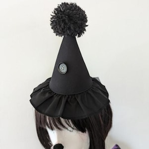 May include: A black cone-shaped hat featuring a ruffled brim, a single black button centered on the front, and a large black yarn pom-pom at the top. The hat is positioned on a mannequin head with dark hair and a black nose accessory.