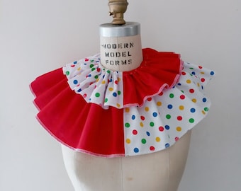 May include: A colorful clown collar with a two-tiered design. The lower tier is solid red, while the upper tier is white with colorful polka dots. The collar is displayed on a mannequin.