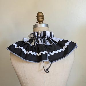 May include: A black and white striped ruffled collar with a wavy white trim. The collar is displayed on a mannequin. The collar has a black tie closure and is made of fabric.