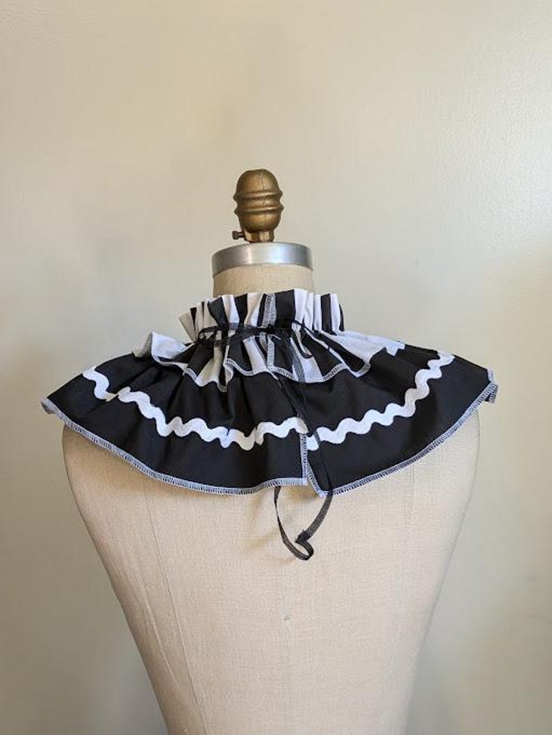 May include: A black and white striped ruffled collar with a wavy white trim. The collar is displayed on a mannequin. The collar has a black tie closure and is made of fabric.