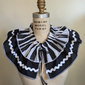 May include: A black and white striped ruffled collar with a scalloped edge, displayed on a mannequin. The collar has a tie closure and is made of fabric. The mannequin has the text "MODERN MODEL FORMS" printed on it.