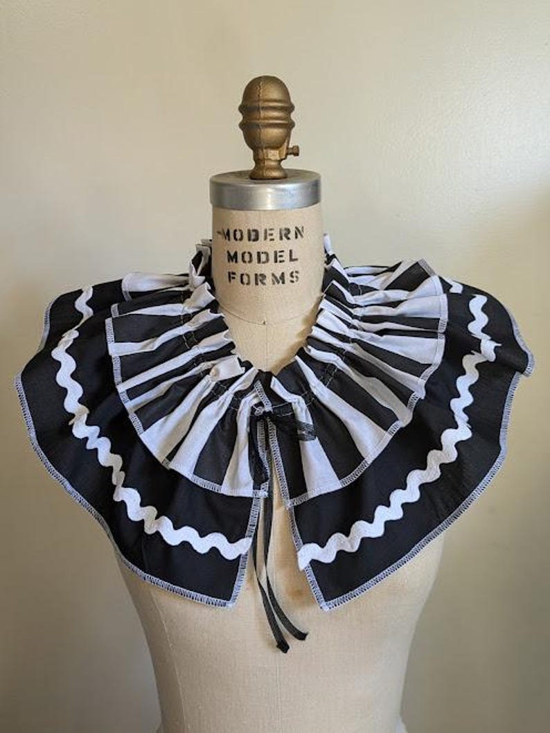 May include: A black and white striped ruffled collar with a scalloped edge, displayed on a mannequin. The collar has a tie closure and is made of fabric. The mannequin has the text "MODERN MODEL FORMS" printed on it.
