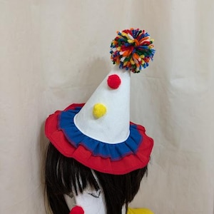 May include: A white cone-shaped hat with a red and blue ruffle trim. The hat has a yellow and red pom-pom on top and a multi-colored pom-pom on the side.