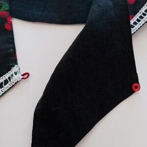 Może przedstawiać: Czarny szalik z tkaniny z tr&oacute;jkątnym zakończeniem, zapinany na czerwony guzik. Szalik ma fragment z wzorem wiśni i białym koronkowym wykończeniem. Szalik jest wykonany z ciemnej tkaniny.