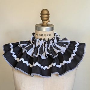 May include: A black and white ruffled collar with a wavy white trim. The collar is on a mannequin with the text "MODERN MODEL" visible. The collar has a vintage aesthetic.