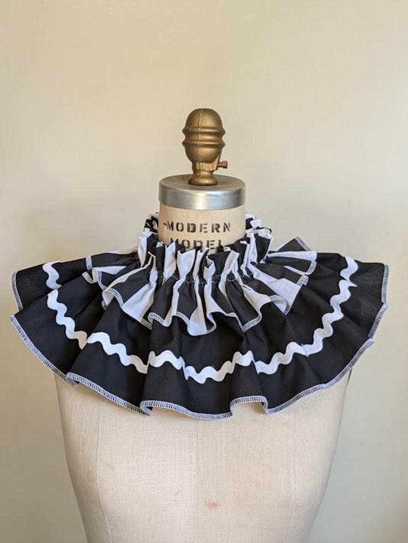 May include: A black and white ruffled collar with a wavy white trim. The collar is on a mannequin with the text "MODERN MODEL" visible. The collar has a vintage aesthetic.