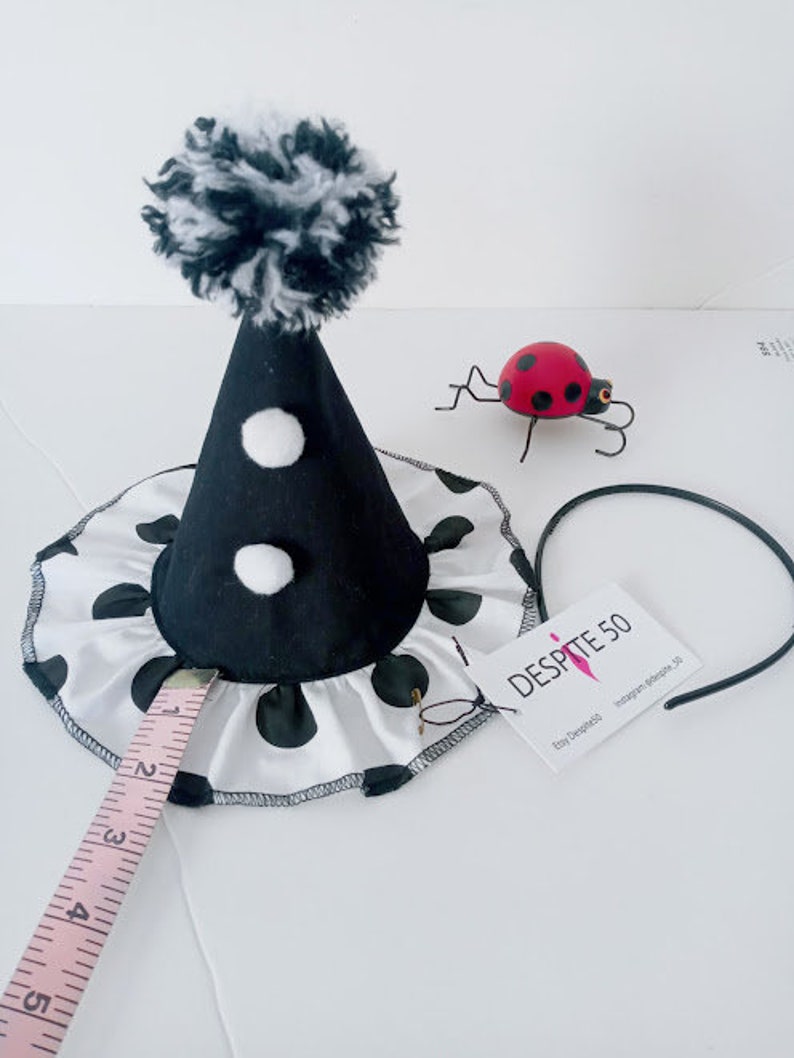 K&ouml;nnte beinhalten: Ein schwarz-wei&szlig; gepunkteter Partyhut mit einem schwarzen Kegel und einem wei&szlig;en Bommel oben. Der Hut liegt auf einer wei&szlig;en Oberfl&auml;che mit einem Ma&szlig;band und einem Marienk&auml;fer.