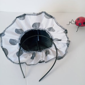 K&ouml;nnte beinhalten: Ein schwarzes Stirnband mit einem wei&szlig;en Stoff-R&uuml;schenbesatz mit schwarzen Punkten. Das Stirnband ist aus einem weichen, flexiblen Material.