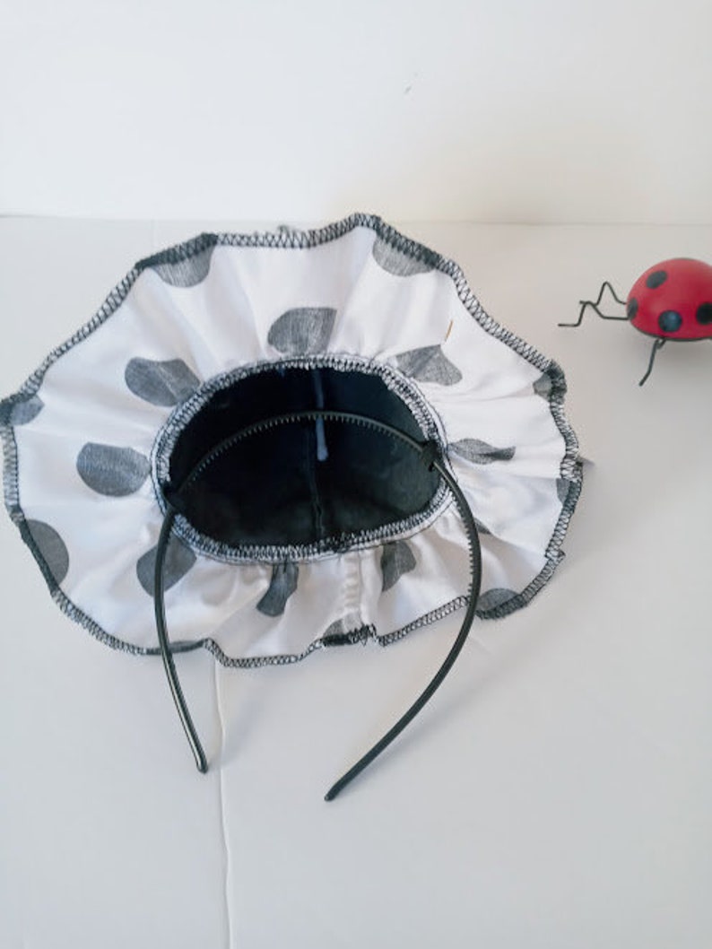 K&ouml;nnte beinhalten: Ein schwarzes Stirnband mit einem wei&szlig;en Stoff-R&uuml;schenbesatz mit schwarzen Punkten. Das Stirnband ist aus einem weichen, flexiblen Material.