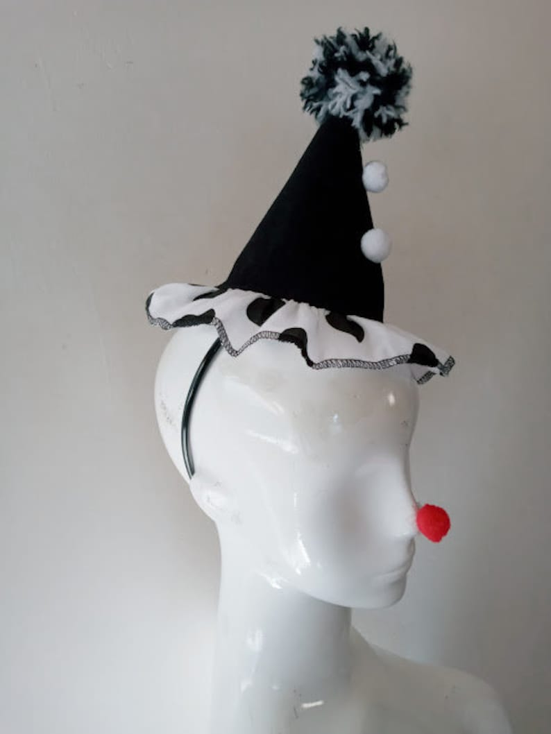K&ouml;nnte beinhalten: Ein schwarz-wei&szlig;er Clownshut mit einem schwarzen Stirnband und wei&szlig;en Bommeln. Der Hut hat einen schwarz-wei&szlig;en Tupfenbesatz.