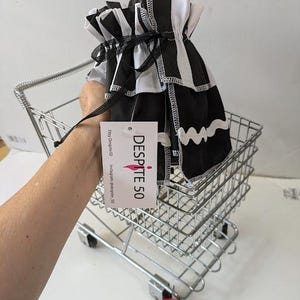 May include: A black and white striped fabric bag with a drawstring closure is held in a small metal shopping cart. The bag has a ruffled top and a wavy white trim. A tag with the text "DESPITE 50" hangs from the bag. A red ladybug figurine is in the foreground.