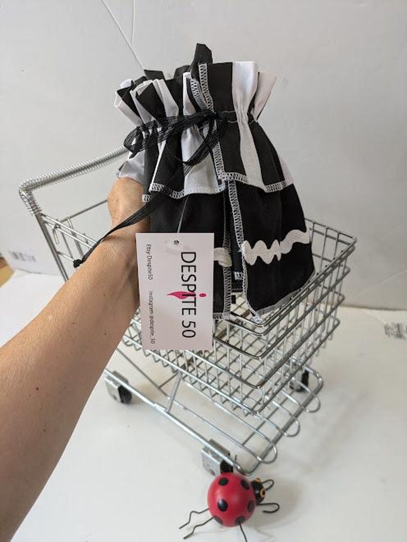 May include: A black and white striped fabric bag with a drawstring closure is held in a small metal shopping cart. The bag has a ruffled top and a wavy white trim. A tag with the text "DESPITE 50" hangs from the bag. A red ladybug figurine is in the foreground.