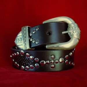 May include: A dark brown leather belt with silver-coloured metal studs and a decorative buckle. The belt features a textured pattern and a cross design made of studs. The buckle has an ornate design.