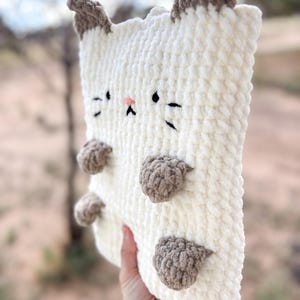 May include: A plush, cream-colored cat-shaped pillow with brown accents. The pillow features embroidered black eyes, whiskers, and a pink nose. Brown paws are attached to the front. The ears are also brown.