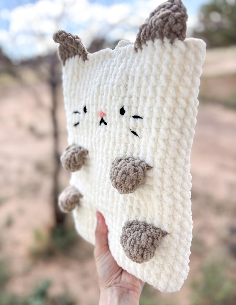May include: A plush, cream-colored cat-shaped pillow with brown accents. The pillow features embroidered black eyes, whiskers, and a pink nose. Brown paws are attached to the front. The ears are also brown.
