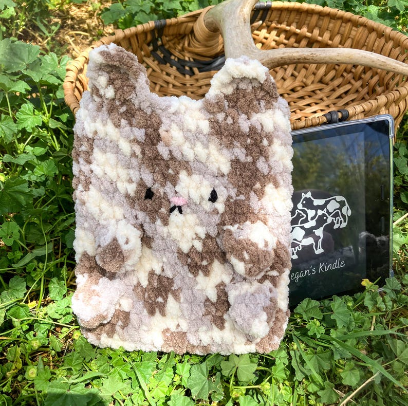 May include: A handmade, plush tablet case in the shape of a cat's head. The case is made of a soft, textured yarn in shades of brown, cream, and gray. It features small black eyes and a pink nose. The text "Megan's Kindle" is visible on the tablet screen.