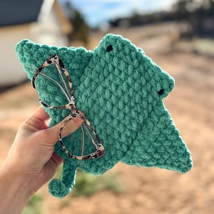 May include: A teal green, crocheted eyeglass case in the shape of a leaf, with two small black button eyes. The case holds a pair of eyeglasses with a tortoiseshell pattern frame.