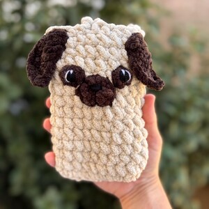 Op de afbeelding: Een handgemaakte, beige gehaakte mopshond met donkerbruine oren, neus en oogdetails. De pluchen hond heeft een rechthoekige vorm. Het gezicht van de hond is gedetailleerd met zwarte ogen en een kleine, donkerbruine neus.