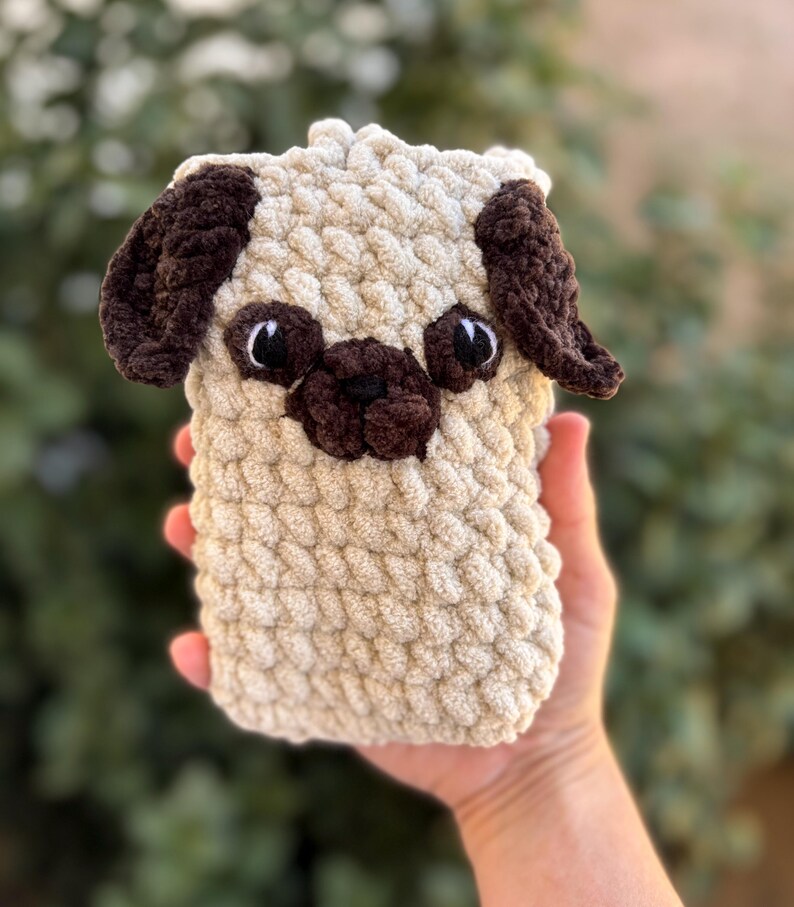 Op de afbeelding: Een handgemaakte, beige gehaakte mopshond met donkerbruine oren, neus en oogdetails. De pluchen hond heeft een rechthoekige vorm. Het gezicht van de hond is gedetailleerd met zwarte ogen en een kleine, donkerbruine neus.