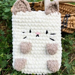 May include: A white, plush, crocheted cat-shaped case with brown ears, paws, and facial details. The case has a rectangular shape and is designed to hold a tablet or other electronic device. The cat's face features black whiskers and a pink nose.