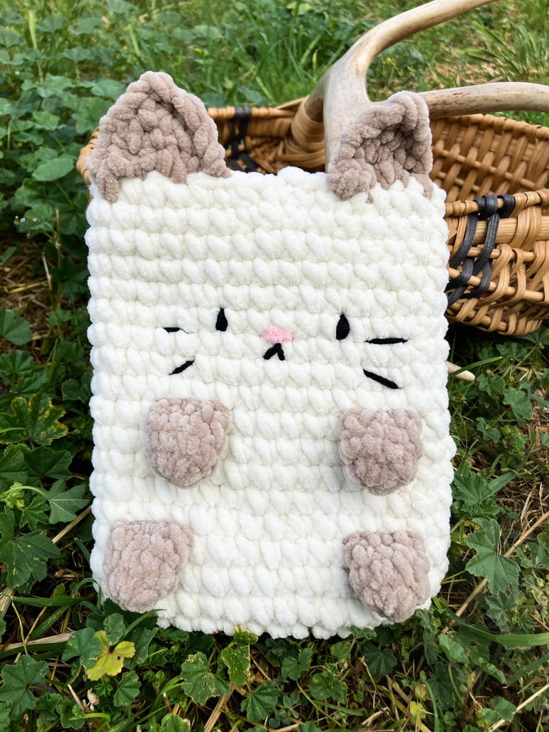 May include: A white, plush, crocheted cat-shaped case with brown ears, paws, and facial details. The case has a rectangular shape and is designed to hold a tablet or other electronic device. The cat's face features black whiskers and a pink nose.