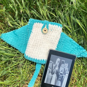 Op de afbeelding: Een turquoise blauwe gehaakte e-reader hoes met een wit midden en een houten knoop. De hoes is manta-vormig en heeft een staart. Een zwart-witte e-reader met het woord "kindle" op het scherm zit in de hoes.