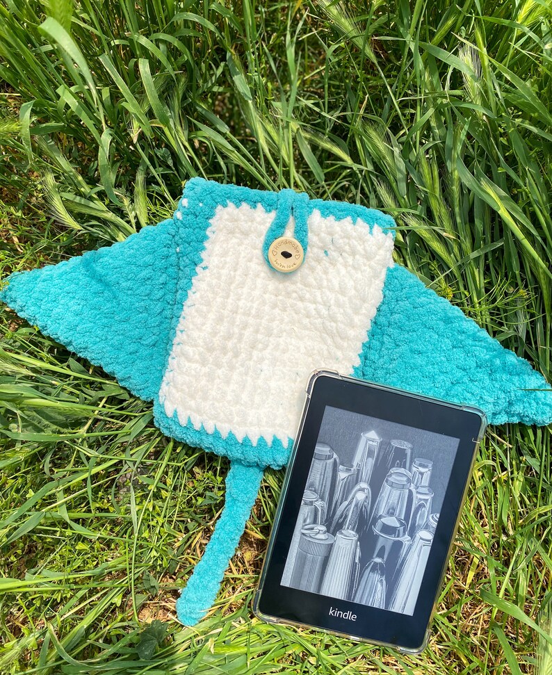 Op de afbeelding: Een turquoise blauwe gehaakte e-reader hoes met een wit midden en een houten knoop. De hoes is manta-vormig en heeft een staart. Een zwart-witte e-reader met het woord "kindle" op het scherm zit in de hoes.