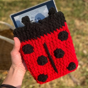 May include: A red and black crocheted ladybug-themed e-reader case. The case features large black spots, a black stripe down the center, and a black top. The e-reader is visible inside the case, displaying text from a book.